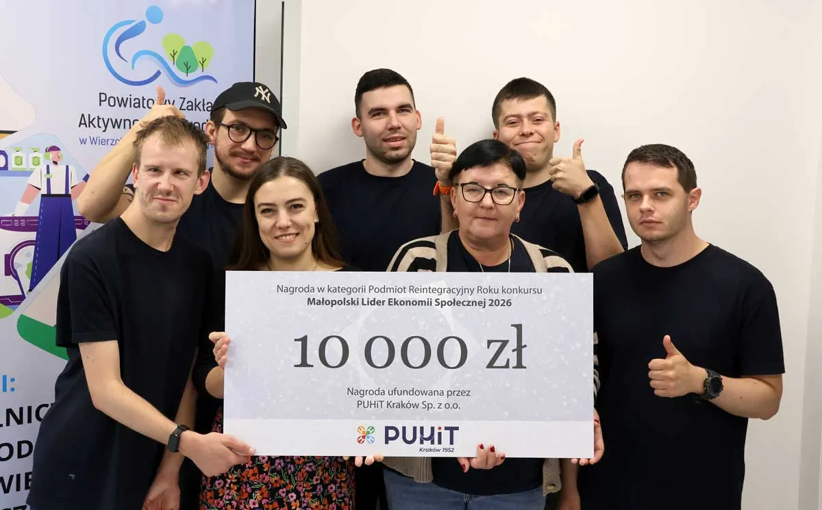 Siedmioro uśmiechniętych pracowników PZAZ przy roll-upie Zakładu trzyma symboliczny czek na 10 000 zł, kilka osób pokazuje kciuki w górę