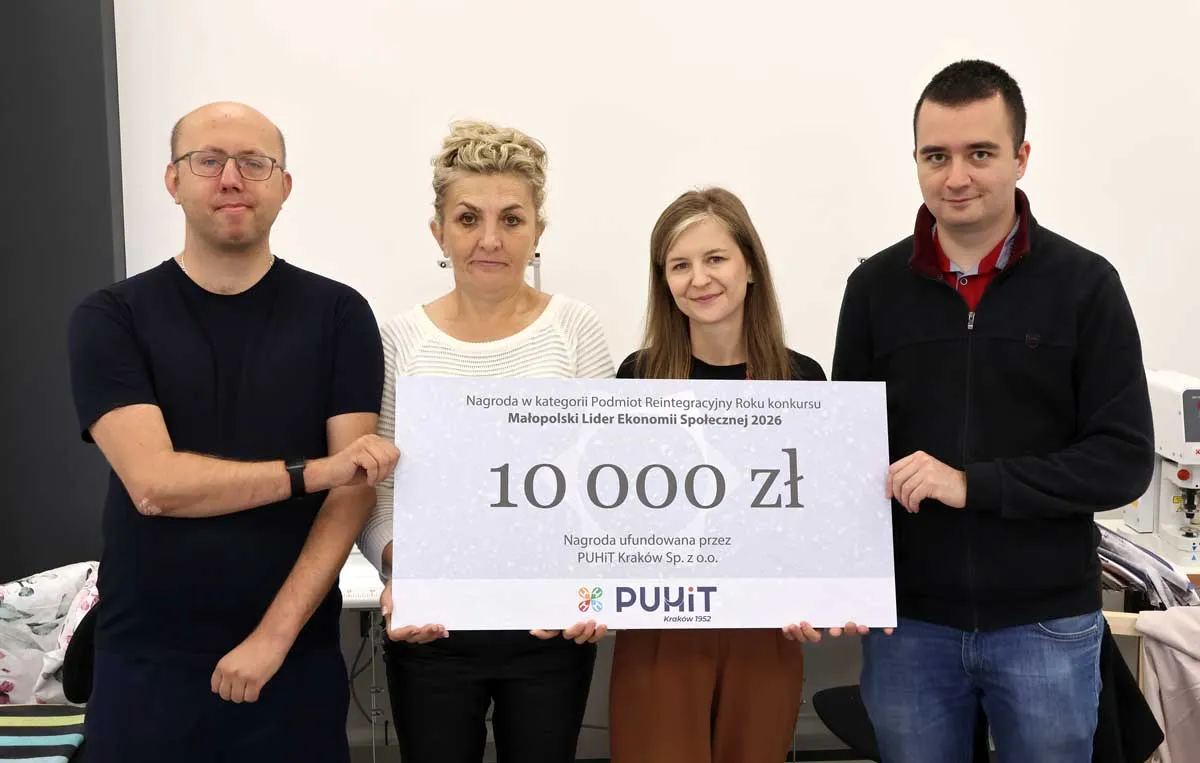 Czworo pracowników PZAZ z symbolicznym czekiem na 10 000 zł w pracowni krawieckiej, w tle maszyna do szycia